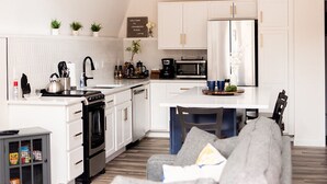 A modern kitchen with white cabinetry, a central island, and a dining area with a grey sofa. - Ridge Point | Walk to Glen Arbor | Pet-Friendly | Close to Sleeping Bear Dunes (Glen Arbor)