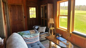 A cozy cabin interior with a leather sofa, a small table, and a view of a green field through large windows. - Rustic Log Cabin with Pond and Breathtaking View (Ossian, NY)