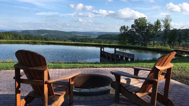 A wooden deck with Adirondack chairs and a fire pit overlooking a lake and distant hills. - Rustic Log Cabin with Pond and Breathtaking View (Ossian, NY)