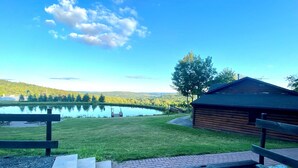 A wooden cabin with a gravel path leading to a dock over a lake, surrounded by greenery and a clear sky. - Rustic Log Cabin with Pond and Breathtaking View (Ossian, NY)