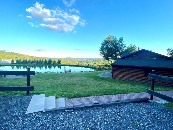 Eine Holzhütte mit einem Kiesweg, der zu einem Steg über einem See führt, umgeben von viel Grün und einem klaren Himmel.
