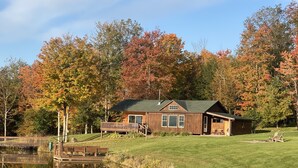 A wooden cabin with a deck and a pond in front. - Rustic Log Cabin with Pond and Breathtaking View (Ossian, NY)