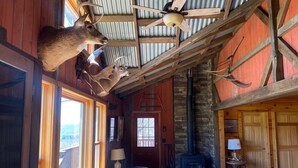 A rustic cabin interior with a stone fireplace, wooden ceiling beams, and deer antlers mounted on the wall. - Rustic Log Cabin with Pond and Breathtaking View (Ossian, NY)