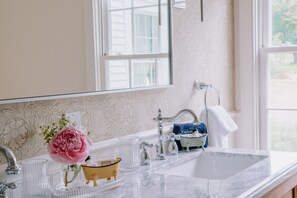 A bathroom with a marble sink, a mirror, a window, and a decorative vase with flowers. - The Millbrook Inn (Millbrook)