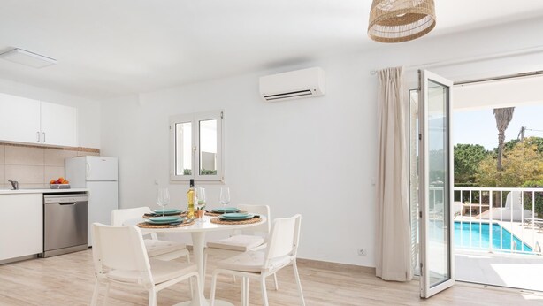 Una cocina moderna con gabinetes blancos, lavavajillas integrado y un comedor con mesa redonda y sillas blancas. Se aprecia una ventana grande con vista a una piscina y zonas verdes.
