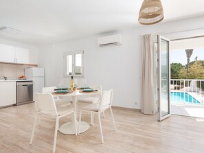 A modern kitchen with white cabinets, a built-in dishwasher, and a dining area with a round table and white chairs. There is a large window with a view of a swimming pool and greenery outside. - New Apartment DOR, Pool, Wifi, Ac, Bbq, Cala Dor (Cala D'or)