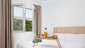 Un dormitorio con cama, ventana con cortinas, mesita de noche con alimentos para el desayuno y vista a la vegetación exterior.