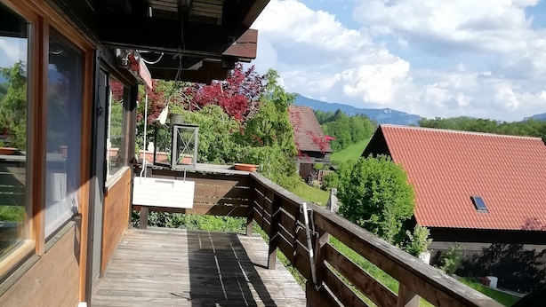 Une terrasse en bois dotée d’une rambarde, offrant une vue sur un paysage rural avec des maisons et des montagnes.