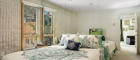 A bedroom with a large bed, a window with blinds, and a door leading to another room.