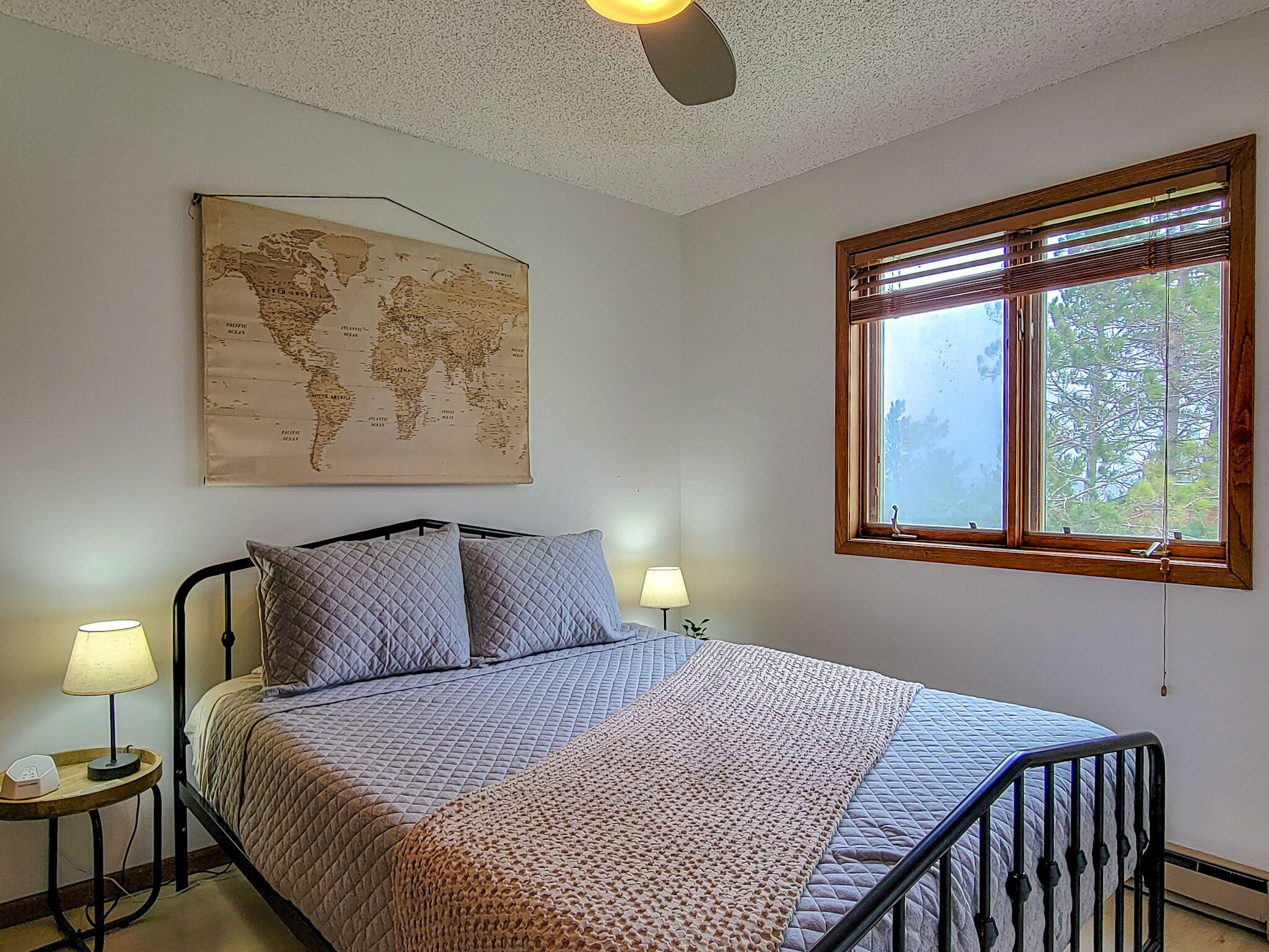 Una cama bien hecha con colcha acolchada y almohadas, un mapa del mundo en la pared, una ventana con persianas y dos lámparas de noche.