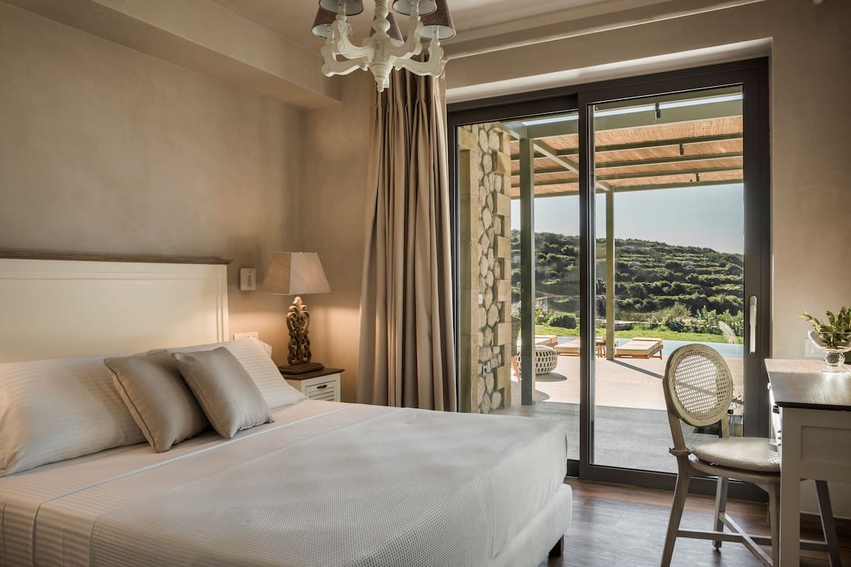 Ein Schlafzimmer mit einem großen Bett, einem Schreibtisch mit Stuhl, einer Lampe und Blick auf einen Garten durch eine Glastür.