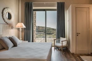 A bedroom with a large bed, a chair, a lamp, and a view of the outdoors through a window. - Zeus Exclusive Villas (Kefalonia)