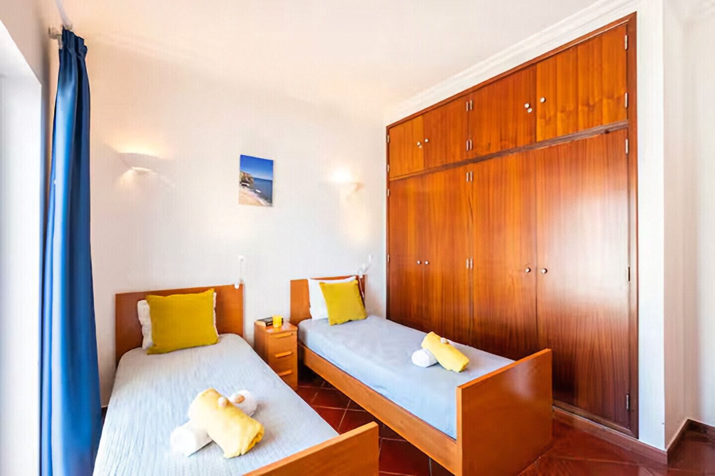 Una camera d'albergo con due letti singoli, un armadio in legno e un tavolino con una lampada.