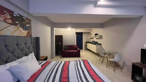 A bedroom with a large bed, a red sofa, a small dining table, and a kitchenette in the background. - Estudio Cozumel A3 (Cozumel)
