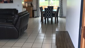 Una sala de estar con un sofá de cuero negro, un banco de madera, una mesa de comedor con sillas estampadas y una cocina al fondo.