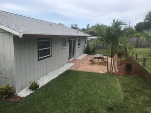 A residential house with a grey exterior, a tiled roof, a small patio with a wooden table and benches, and a well-maintained lawn.