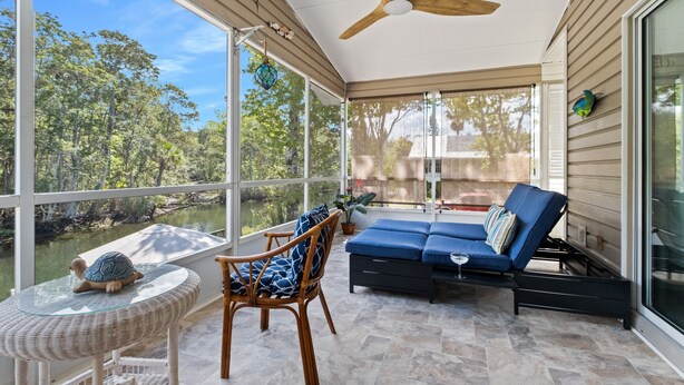 A covered patio with a ceiling fan, wicker furniture, a blue sofa, and a view of a forested area.
