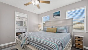 A bedroom with a bed, a ceiling fan, a nightstand, a lamp, a window with blinds, and a wall-mounted shelf.