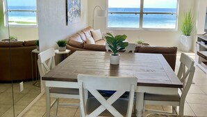 A spacious living area with a dining table, white chairs, a ceiling fan, and a view of the ocean through large windows.