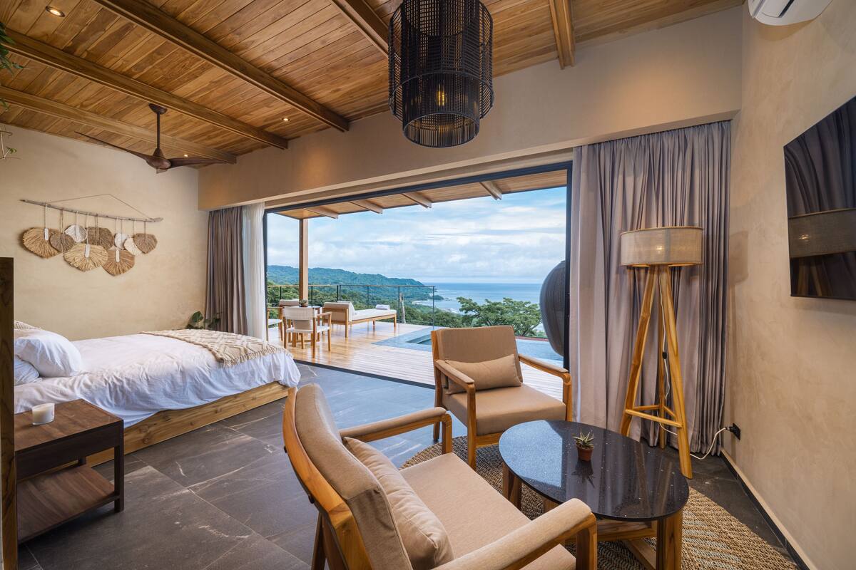 Un dormitorio con cama, una mesita y una silla. Se ve un ventanal con vistas al océano y colinas.