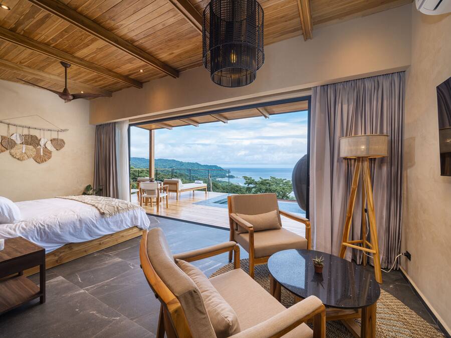 A bedroom with a bed, a small table, and a chair. There is a large window with a view of the ocean and hills.