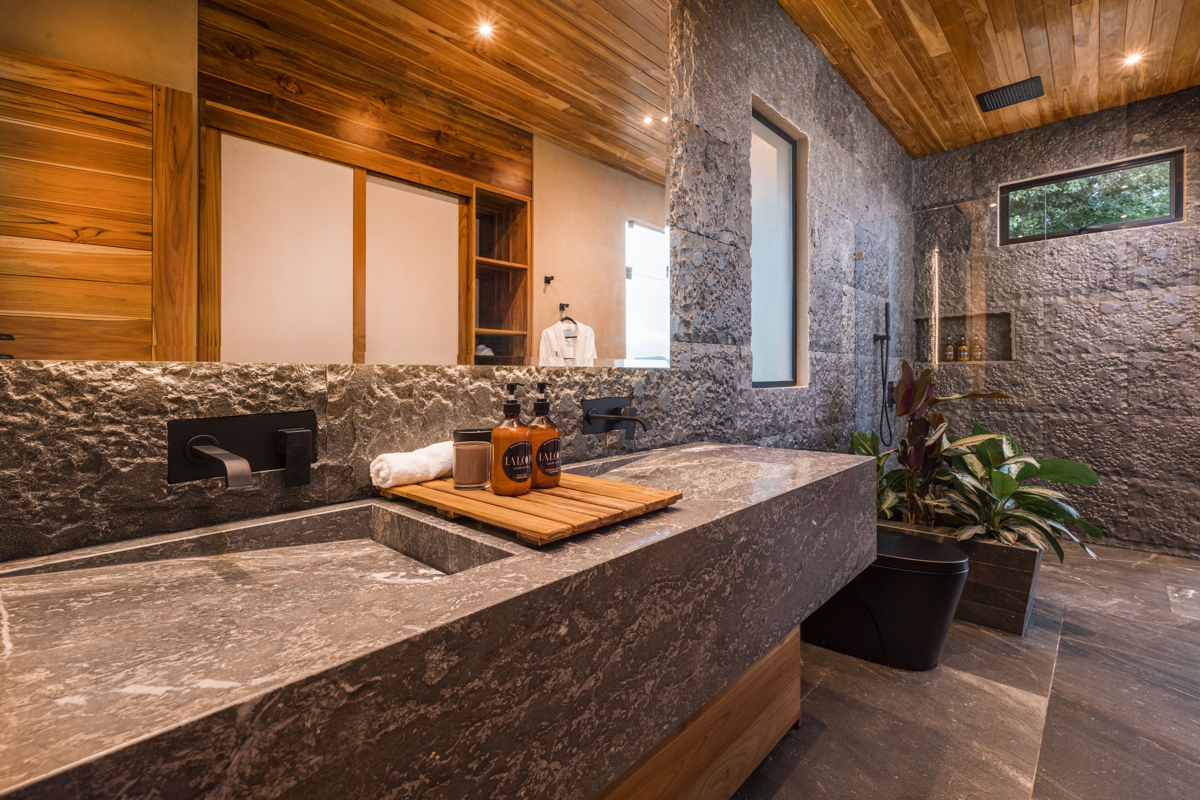 Un baño moderno con pared de piedra, techo de madera y un espejo grande.