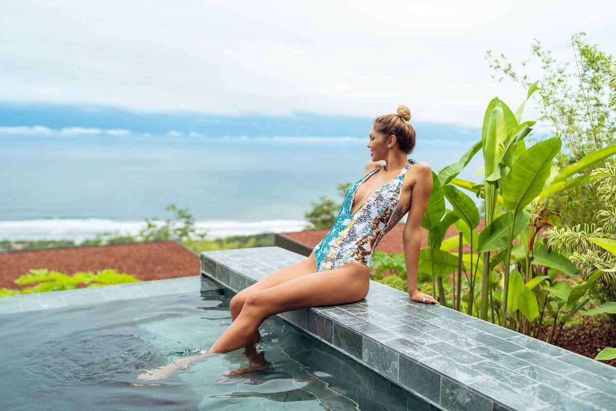 Una mujer sentada en el borde de la piscina con vista al océano.