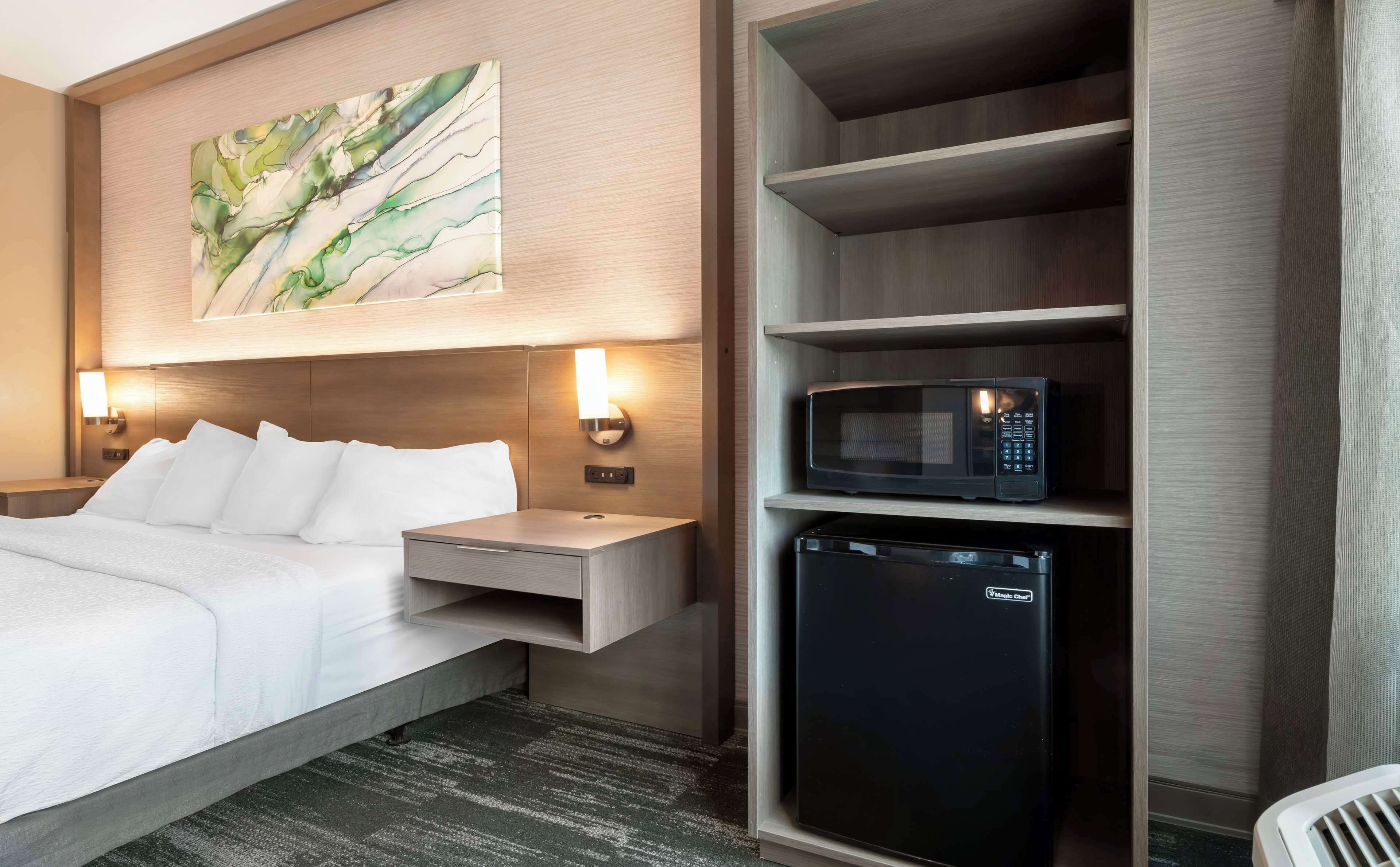 A hotel room with a bed, bedside tables, a wall-mounted light, a microwave, and a refrigerator.