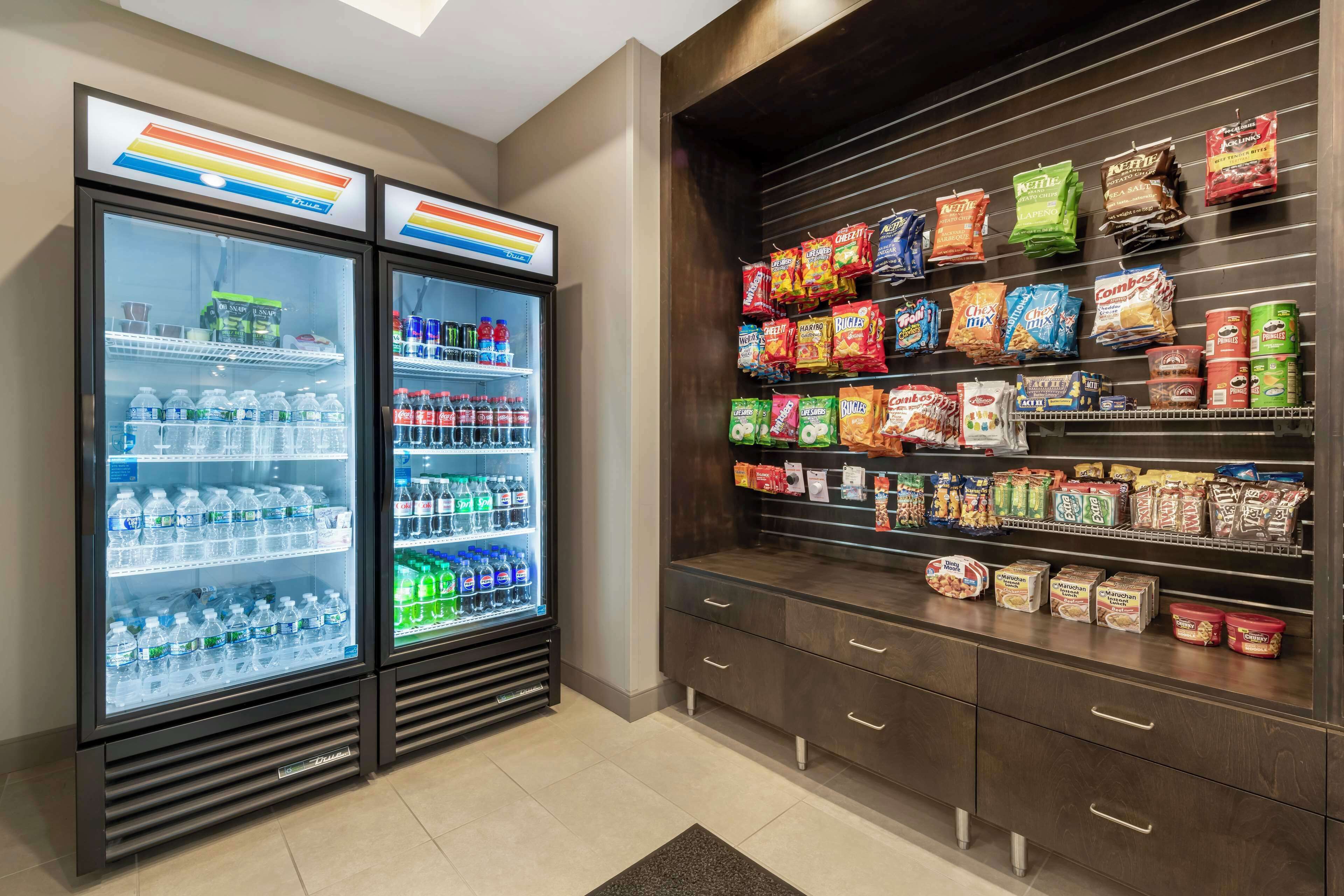 Two refrigerated beverage dispensers stocked with various drinks, a snack display with multiple brands, and a wooden cabinet with drawers.