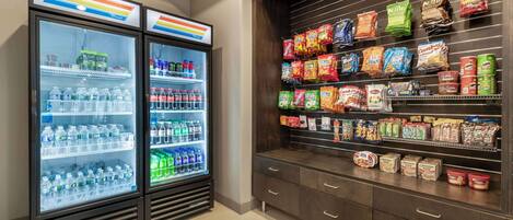 Two refrigerated beverage dispensers stocked with various drinks, a snack display with multiple brands, and a wooden cabinet with drawers.