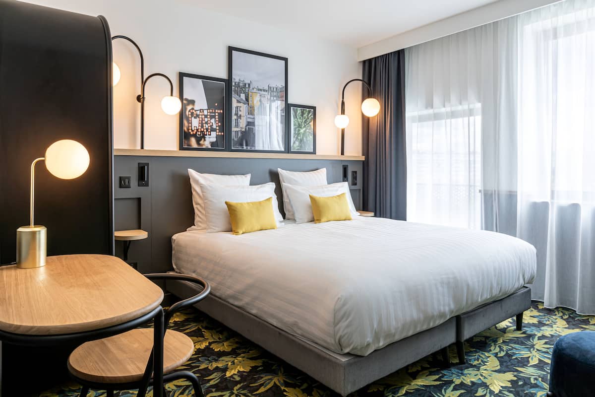 Habitación de hotel con una cama grande, dos mesitas de noche, una mesa redonda de madera y una silla. Hay luces fijadas a la pared y un cuadro enmarcado sobre la cama.