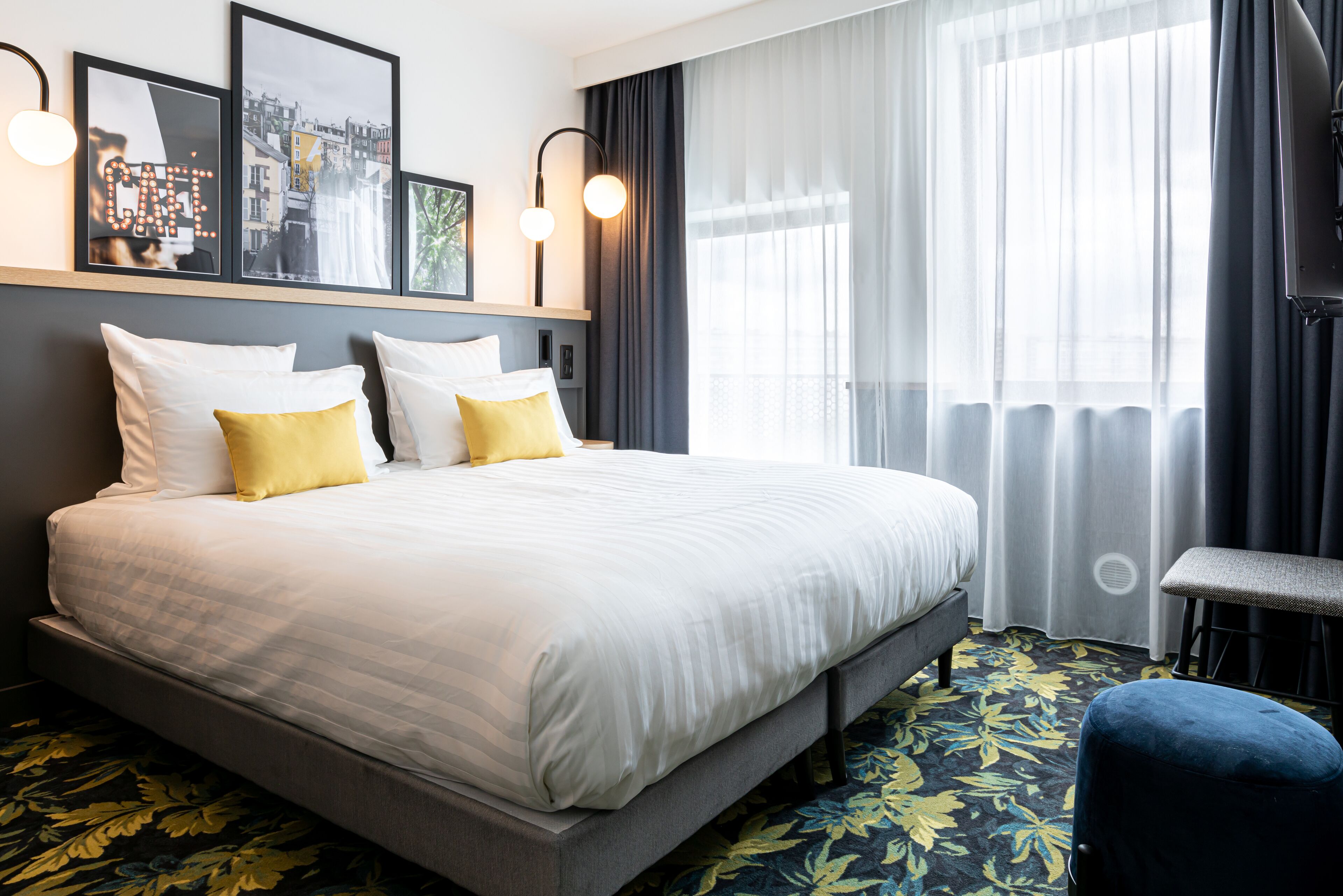 A neatly made bed with white and yellow pillows, a grey headboard, and a floral-patterned carpet.