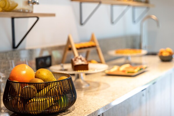 A kitchen counter with a fruit bowl containing oranges and lemons, a loaf of bread, and a plate with pastries. - Best Western M-Treize Paris Asnieres (Asnieres-sur-Seine)