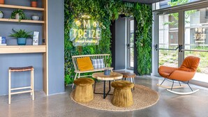 A modern hotel lobby with a green wall, wooden shelves, a seating area with a table and chairs, and a large window.