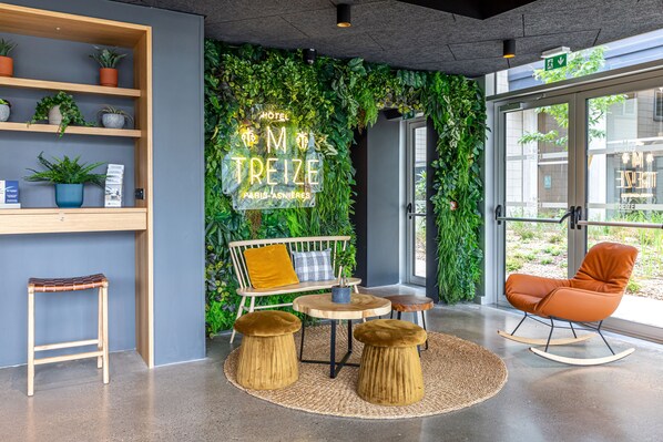 A modern hotel lobby with a green wall, wooden shelves, a seating area with a table and chairs, and a large window. - Best Western M-Treize Paris Asnieres (Asnieres-sur-Seine)