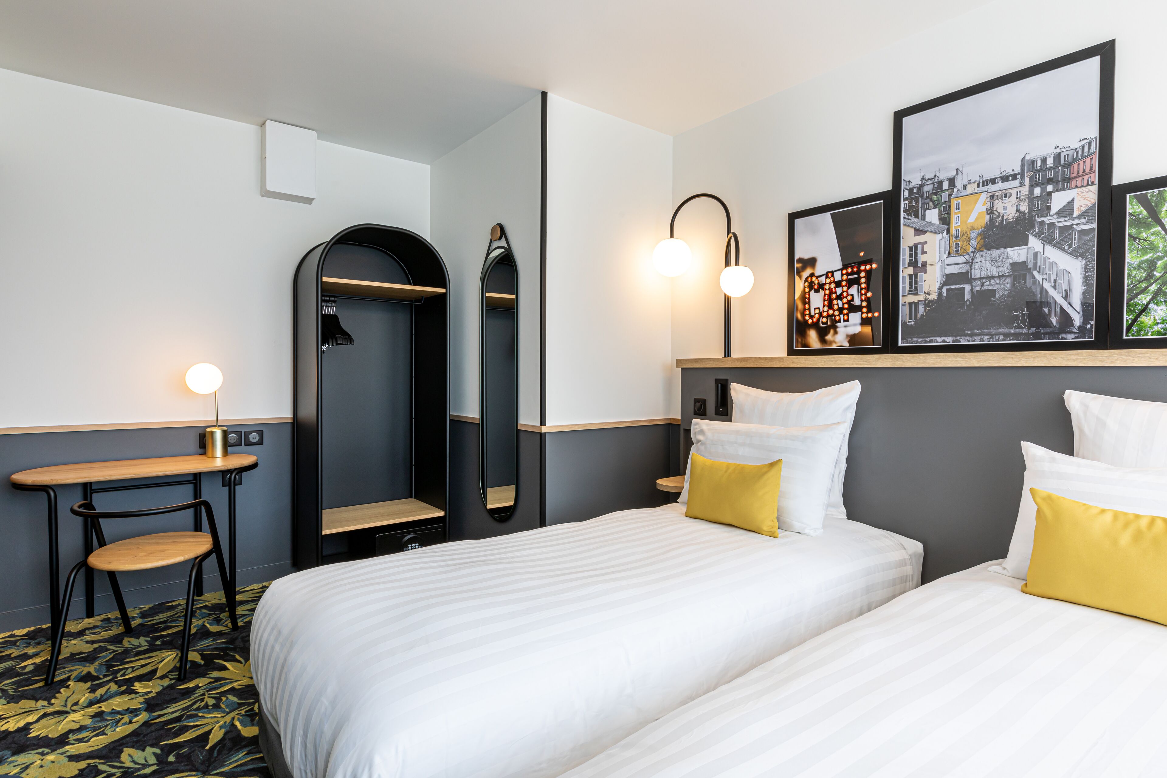 A hotel room with two beds, a mirror, a desk, and a framed cityscape picture on the wall.