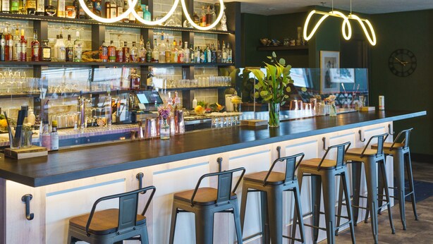 A bar area with a long counter, multiple stools, and shelves stocked with bottles. - The Scout INN (Cody)