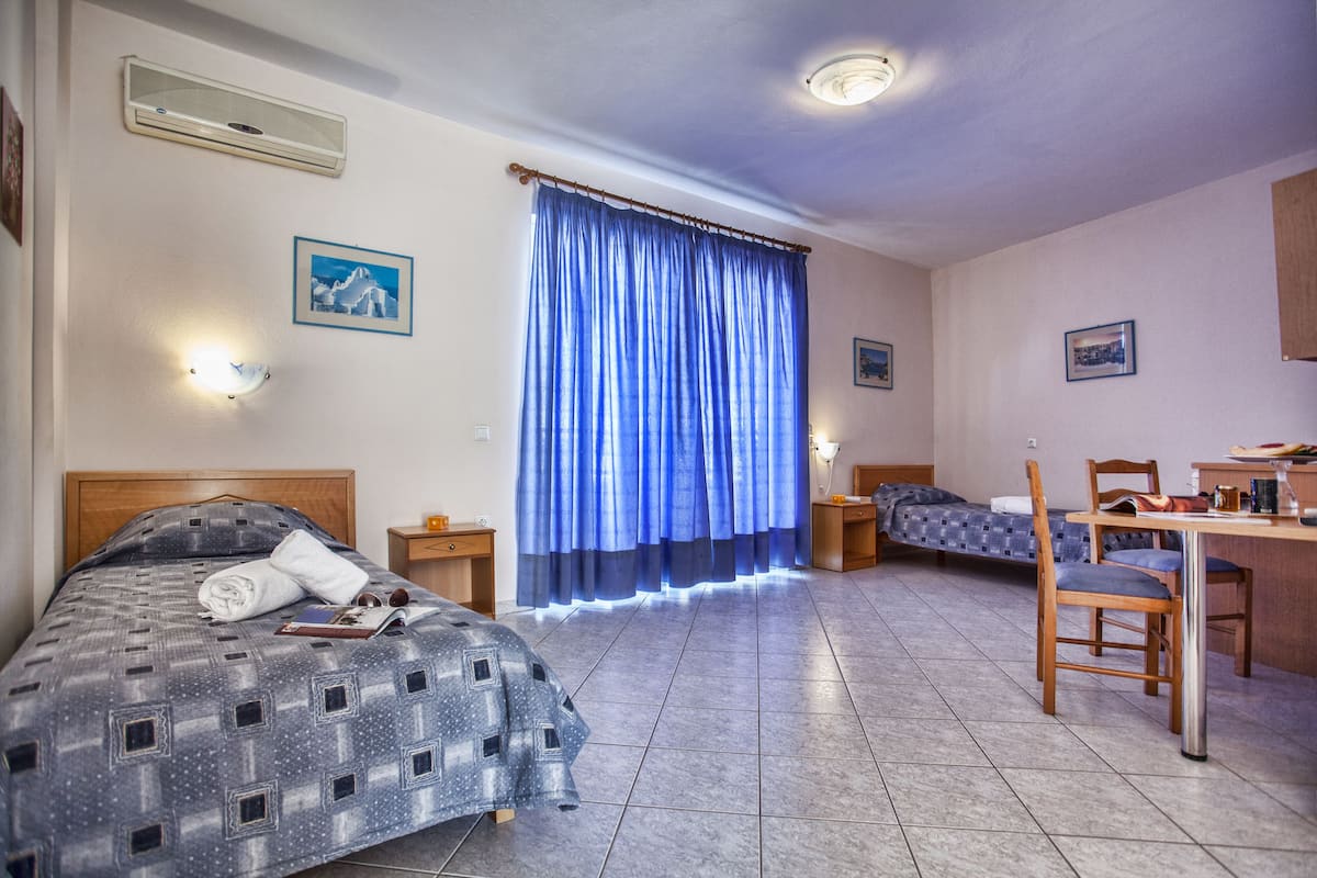 A hotel room with a bed, a desk, a chair, a small table, a window with curtains, and a wall-mounted air conditioner.