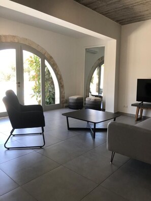 Un salon moderne avec un canapé, une table basse et un fauteuil. Un téléviseur est posé sur un meuble en bois. La pièce est dotée de grandes fenêtres à arcades et offre une vue sur la verdure environnante.