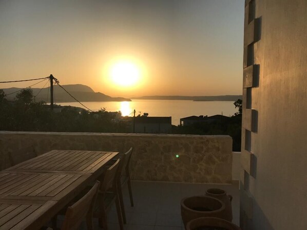Une terrasse avec une table et des chaises en bois, d’où l’on peut admirer un coucher de soleil sur une étendue d’eau, avec des collines lointaines en toile de fond.