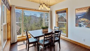 A dining area with a wooden table and chairs, large windows with a view of trees and mountains, and a hanging light fixture. - Modern 3 Bedroom Cabin with Mountain Views! (Sevierville)