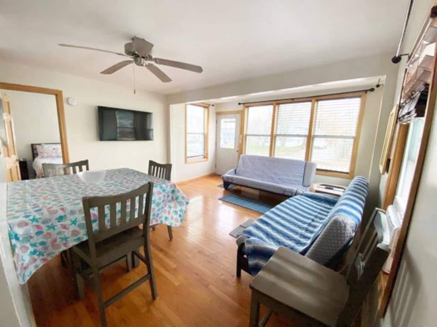 A room with a dining table, chairs, a sofa, a ceiling fan, and large windows.