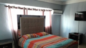 Een slaapkamer met een bed, gordijnen, een airconditioner, een televisie en een nachtkastje.