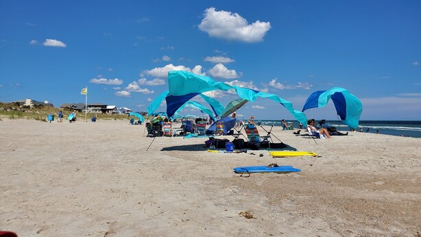 Una playa con gente relajándose bajo toldos azules, algunas sillas y una tabla de surf en la arena.