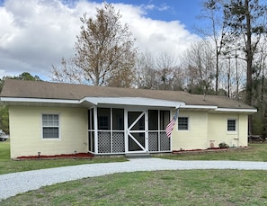 A single-story house with a screened porch, a small front yard with red mulch, and an American flag.