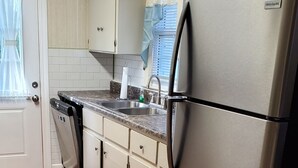 Una cocina con un refrigerador de acero inoxidable, lavavajillas y un lavamanos con grifo. Hay armarios y una ventana con cortina.