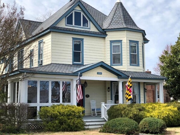 A two-story house with a gabled roof, a porch with a white railing, and two flags on the front.