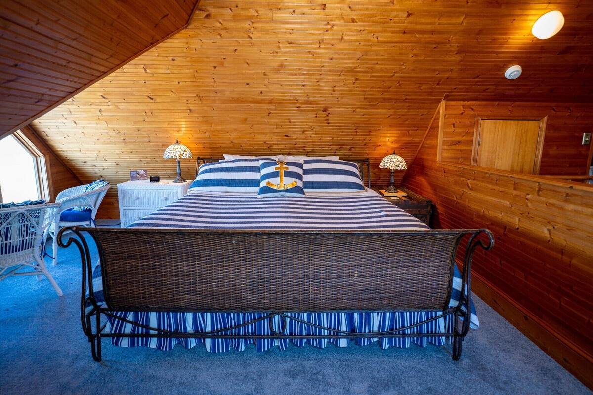 Quarto de cabana de madeira acolhedor, com uma cama grande, duas mesas de cabeceira, uma cadeira de vime e uma janela.