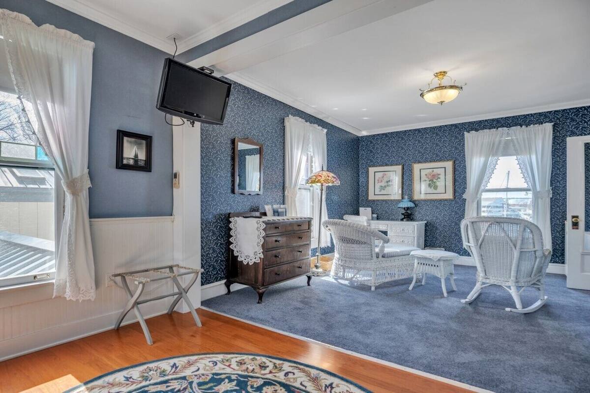 Quarto com esquema de cores azul e branco, com piso de madeira, uma cadeira de balanço branca, cômoda e uma televisão fixada na parede.