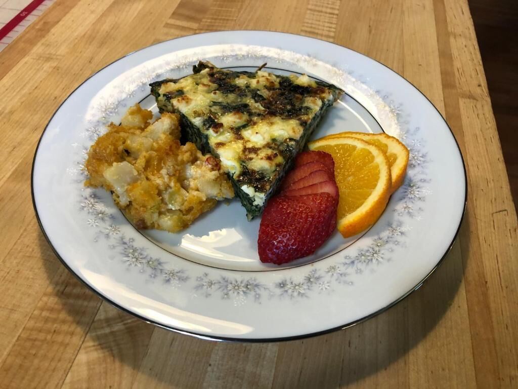 A plate with a slice of quiche, a side of potato salad, a strawberry, and an orange slice on a floral-patterned plate.